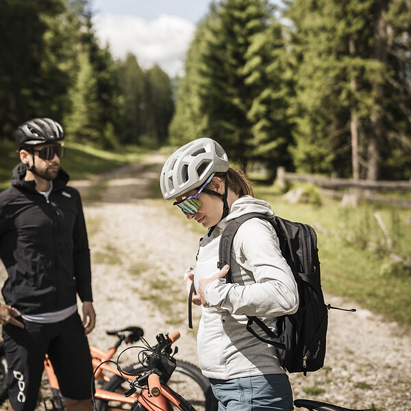 Zwei Mountainbiker mit Fahrradhelmen und ihren Fahrrädern auf einem Weg zwischen hohen Nadelbäumen.