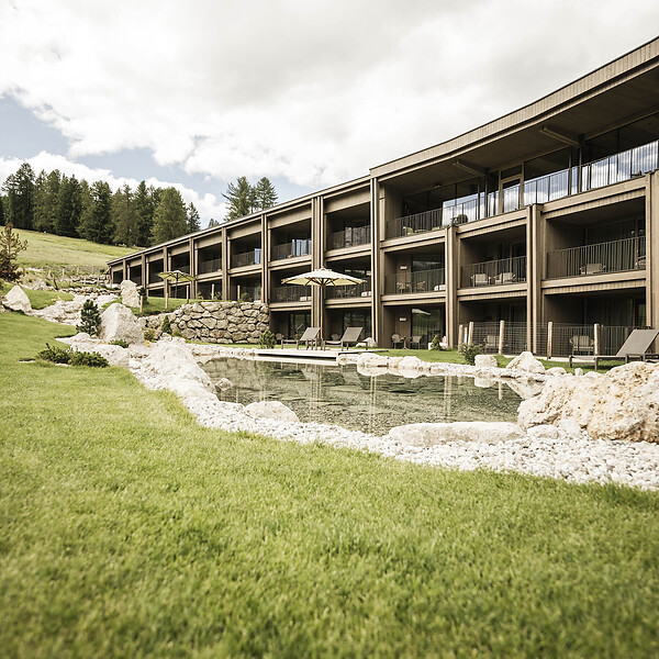 Außenansicht des Hotel Cendevaves mit großzügigen Holzbalkonen, Natursteingarten und grüner Liegewiese vor bewaldeten Berghängen.