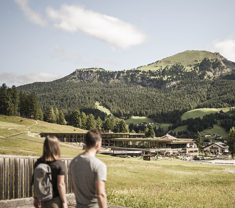 Zwei Personen von hinten an einem Aussichtspunkt mit Holzgeländer und Blick über grüne Bergwiesen und bewaldete Berge.
